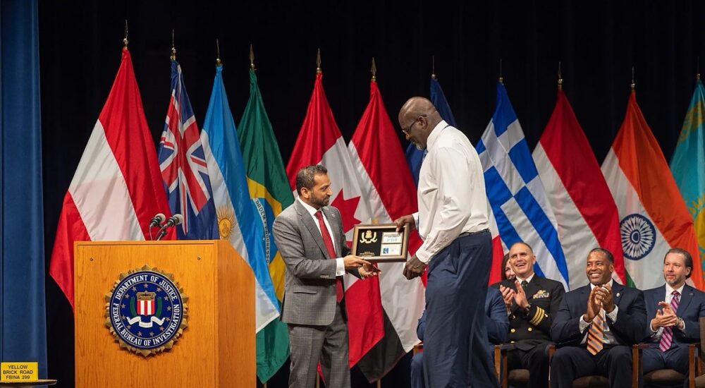 Shaquille O'Neal accepts a framed plaque on stage at an FBI ceremony as seated officials applaud.