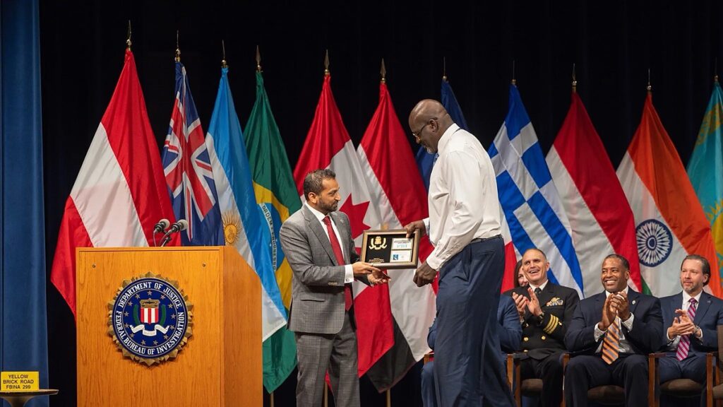 Shaquille O'Neal accepts a framed plaque on stage at an FBI ceremony as seated officials applaud.