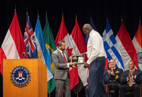 Shaquille O'Neal accepts a framed plaque on stage at an FBI ceremony as seated officials applaud.